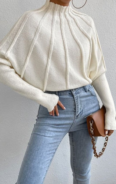 Person wearing a cream sweater and light blue jeans with a brown handbag on a plain background