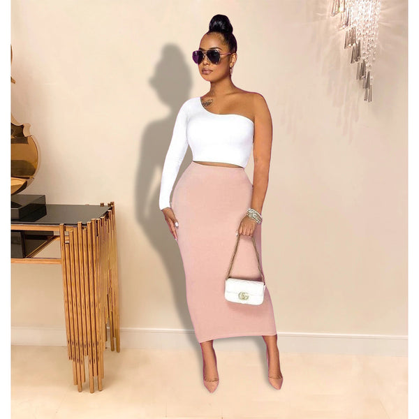 Woman in a white top and pink skirt standing indoors with a side table and decorative items in the background.