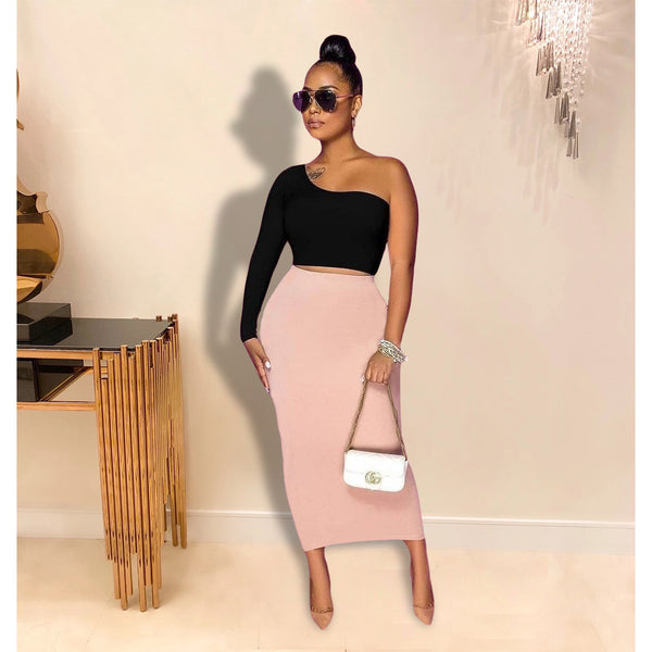 Woman in black one-shoulder top and pink skirt holding a white handbag indoors.