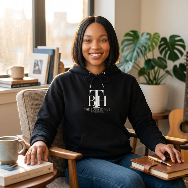SlayEdgeBoutique black pullover hoodie with The Blessed Hub International logo worn by a smiling woman in a home office
