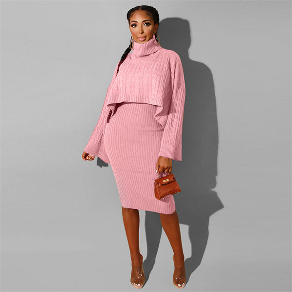 A woman models a pink ribbed knit two-piece set consisting of a cropped turtleneck sweater and a matching knee-length dress, paired with a small brown handbag and clear high-heeled sandals, set against a plain gray background.